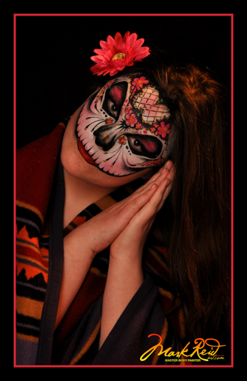 woman with a stylized skull mask painted starting at her upper lip and up. pink highlights match the flower behind her ear